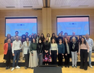 Grupo de participantes del consorcio TRANSIS LAB posando en un auditorio durante la tercera reunión del proyecto, delante de dos pantallas con la imagen corporativa del programa.